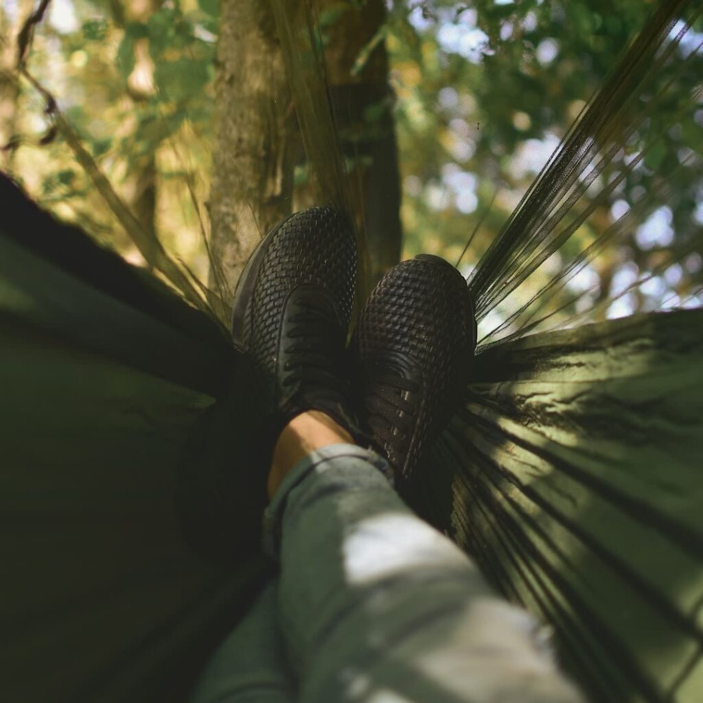 A person relaxes in a hammock tied to a tree, with legs outstretched and feet in dark shoes, surrounded by a lush, green forest.